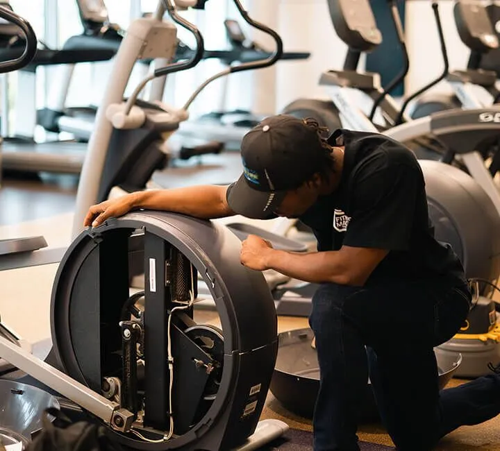 A technician in a black cap inspects the internal mechanics of a fitness machine at a gym