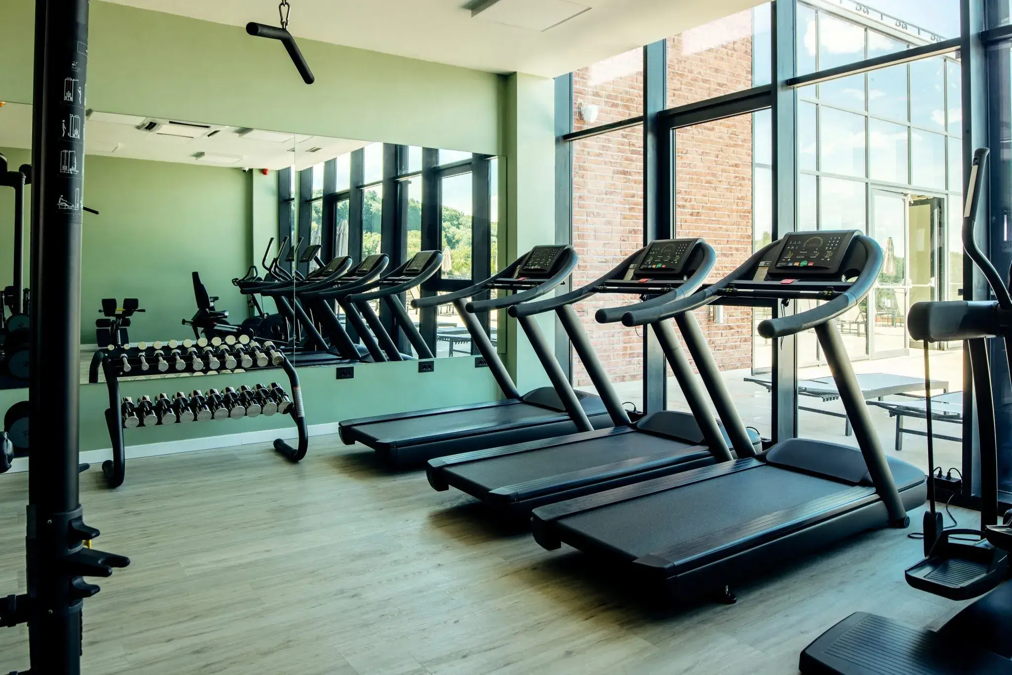 A bright, modern gym features multiple empty treadmills, large mirrors, and expansive windows